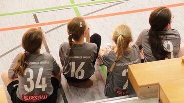 Vier junge Handballspielerinnen einer Jugendmannschaft sitzen in grauen Trikots auf der Bank in der Sporthalle, Rückansicht während eines Jugendhandballspiels